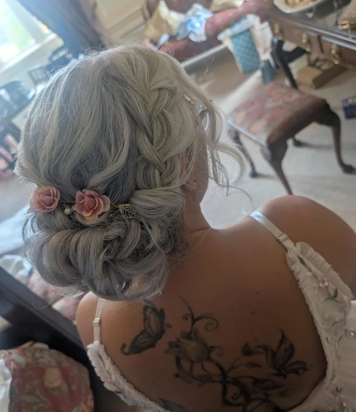 A silver braided low bun finished with small pink flowers and pearl details.
